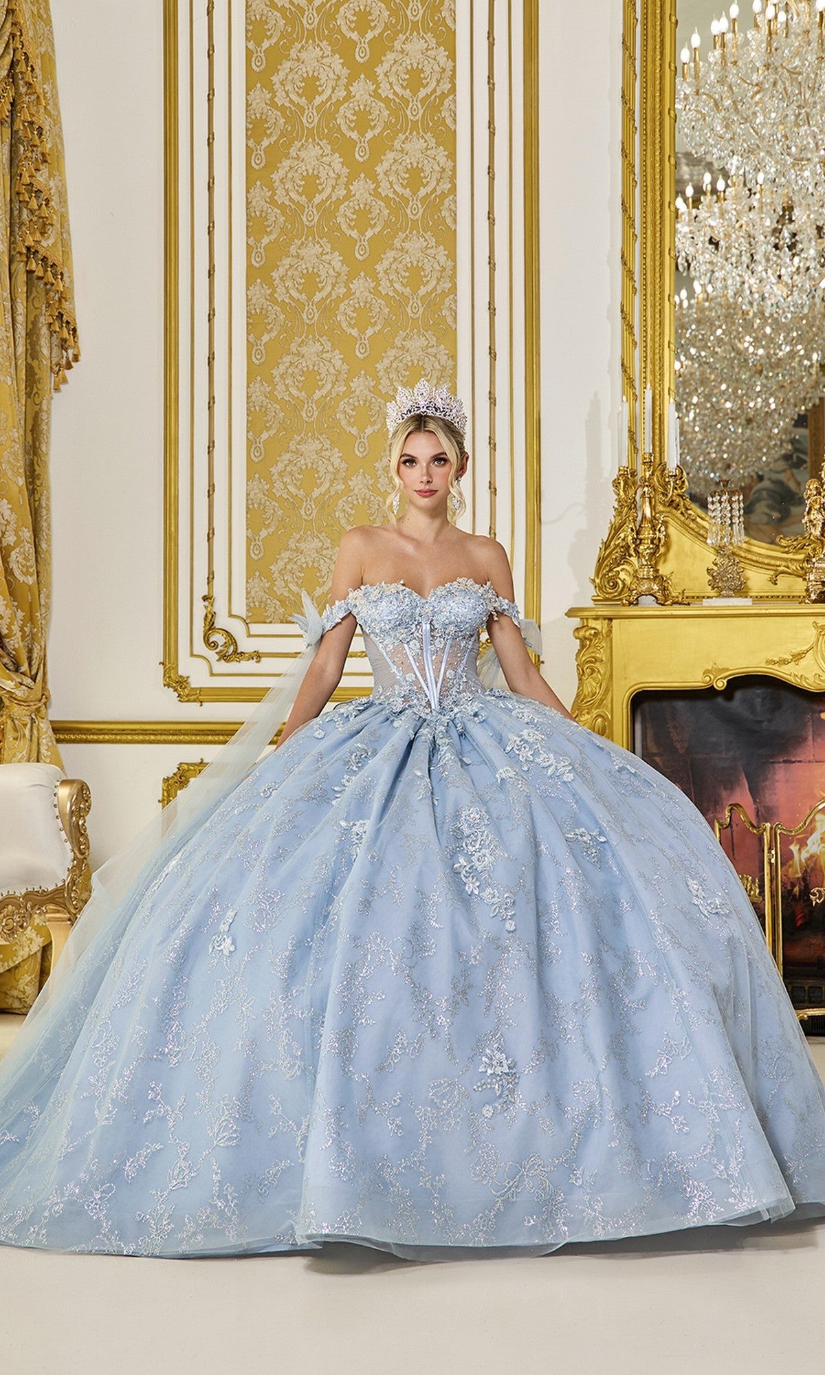 A woman in the Quinceanera Dress 5074 by Dancing Queen, a dusty blue ball gown, stands in an ornate, gold-accented room with a fireplace and chandelier, wearing a tiara.